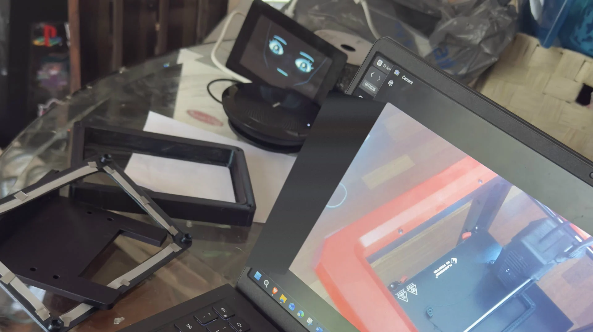 Unassembled enclosure parts on a glass table beside the running prototype, with a laptop showing the printer that is making them.